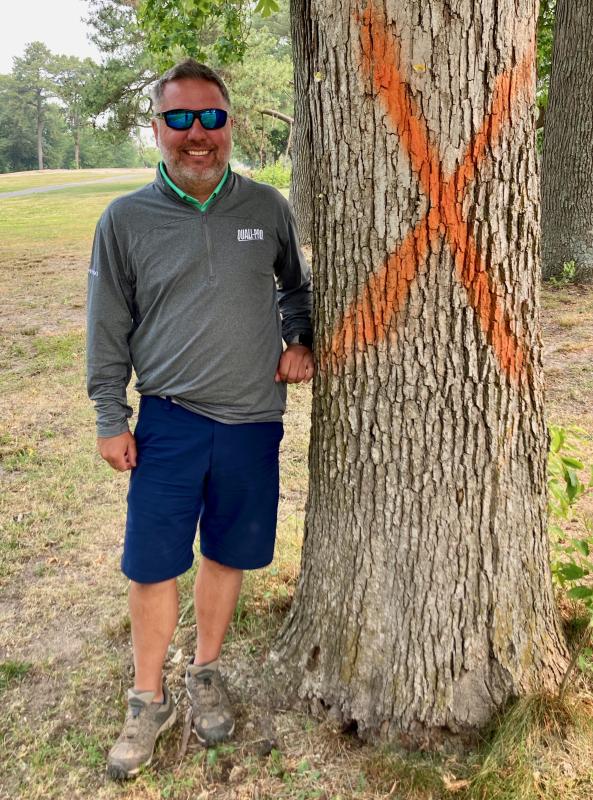 Southern Delaware Golf Club’s Bill Kupfer stands next to a tree on the property that is marked X for removal. Hundreds of similar trees have already been cut down, while others will be limbed up to provide light and air for nearby turf. This tree stands (for now) between the 12th green and the tee box for the 16th hole.